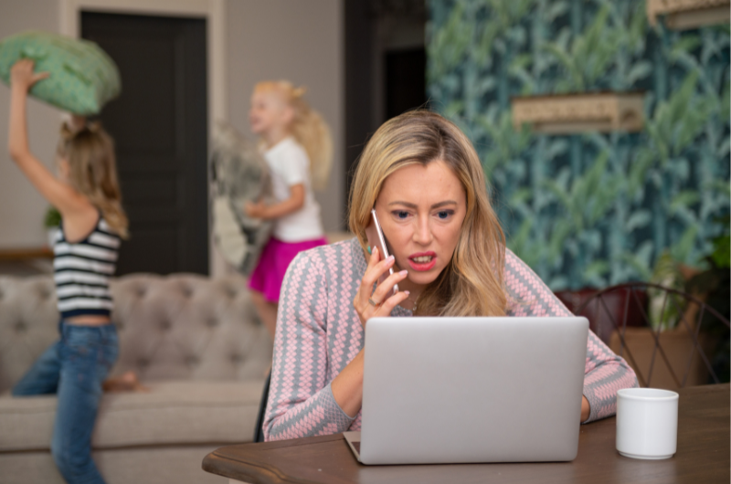 Women with blonde hair, looking stressed, on the phone, while looking at laptop. Children playing in the background making a mess