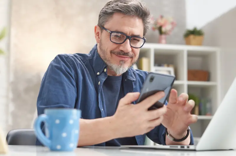 Man in his 40s wearing glasses smiles while using a smartphone at a desk, with a laptop and a blue mug nearby in a bright home office setting.