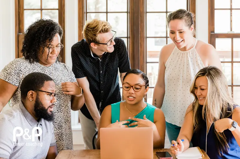 A group of six colleagues from diverse backgrounds collaborate around a laptop, discussing ideas and working together in a bright office setting. The image represents career coaching, teamwork and professional development.