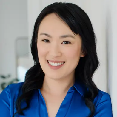 Professional headshot of a smiling woman (our Canadian www.parentandprofessional.co.uk 1 Leadership development coach Joanna Li) with long dark hair, wearing a bright blue blouse. She is standing indoors against a light, neutral background, looking directly at the camera with a warm and approachable expression. The image conveys confidence, professionalism, and friendliness.